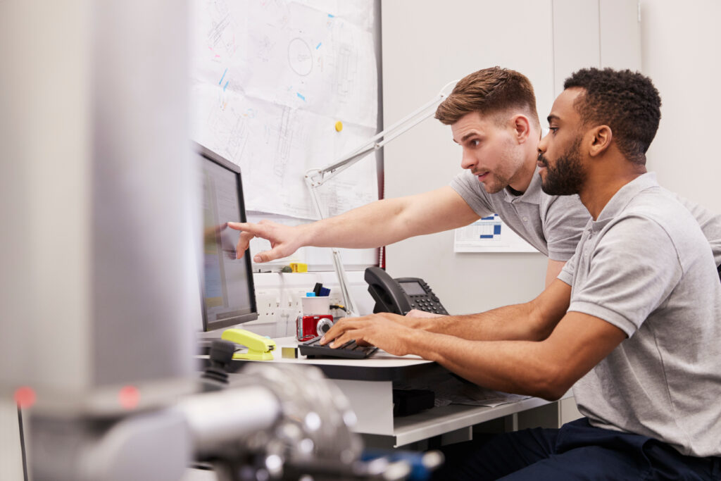 Two male engineers analyzing data on a computer screen using a coordinate measuring machine (CMM) in a factory setting.