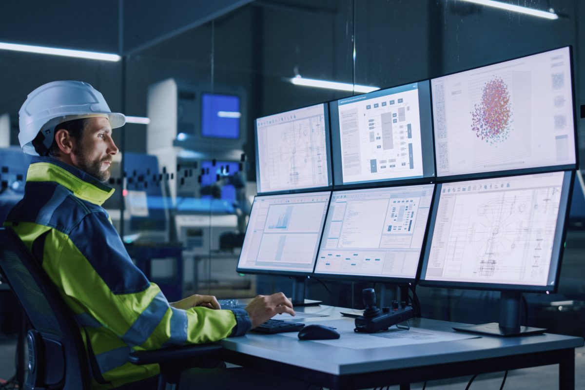 Industry 4.0 Modern Factory: Facility Operator Controls Workshop Production Line, Uses Computer with Screens Showing Complex UI of Machine Operation Processes, Controllers, Machinery Blueprints.